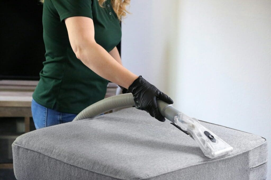 a woman in a green shirt and black gloves vacuuming a gray ottoman