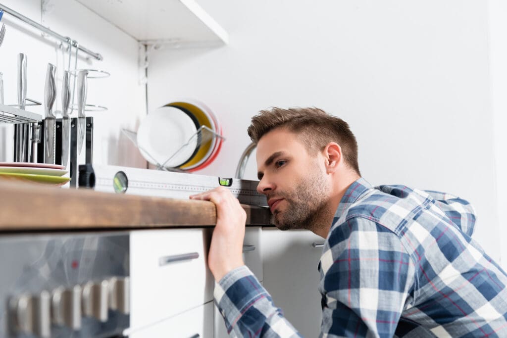 concentrated young man looking at measuring level tool on table on blurred foreground in kitchen