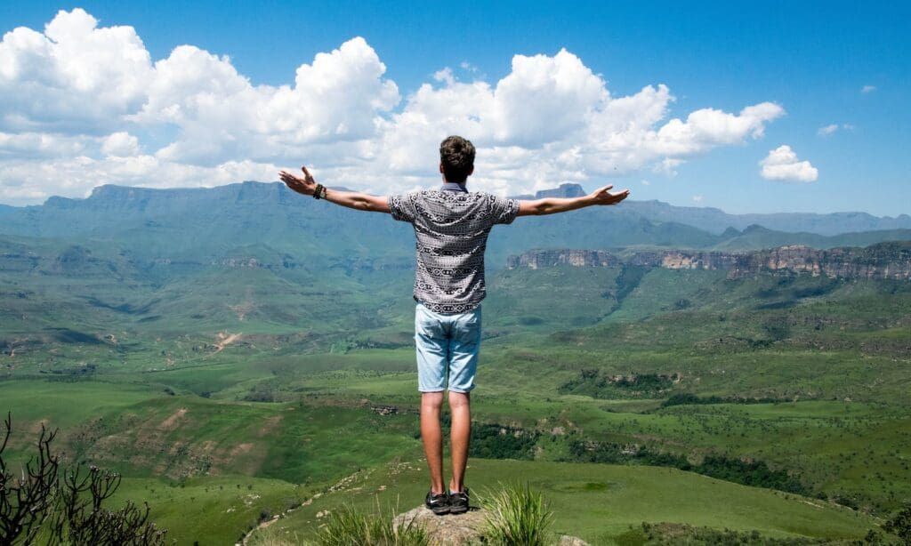 A man stands triumphantly with arms open, overlooking a scenic mountain range.