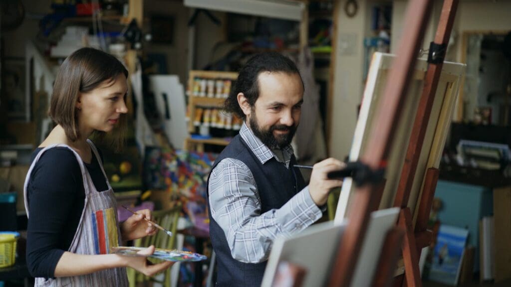 Man painting on canvas with woman assisting in a creative studio setting.