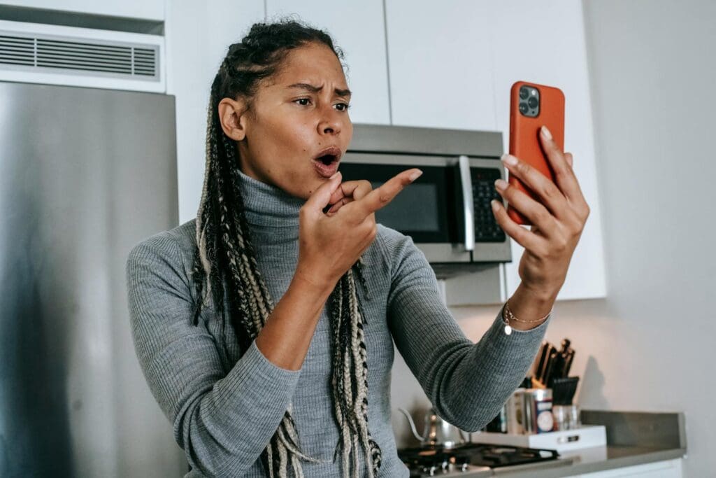 Angry young African American female with long braids in casual clothes pointing at screen nervously while disputing during video call on mobile phone in kitchen