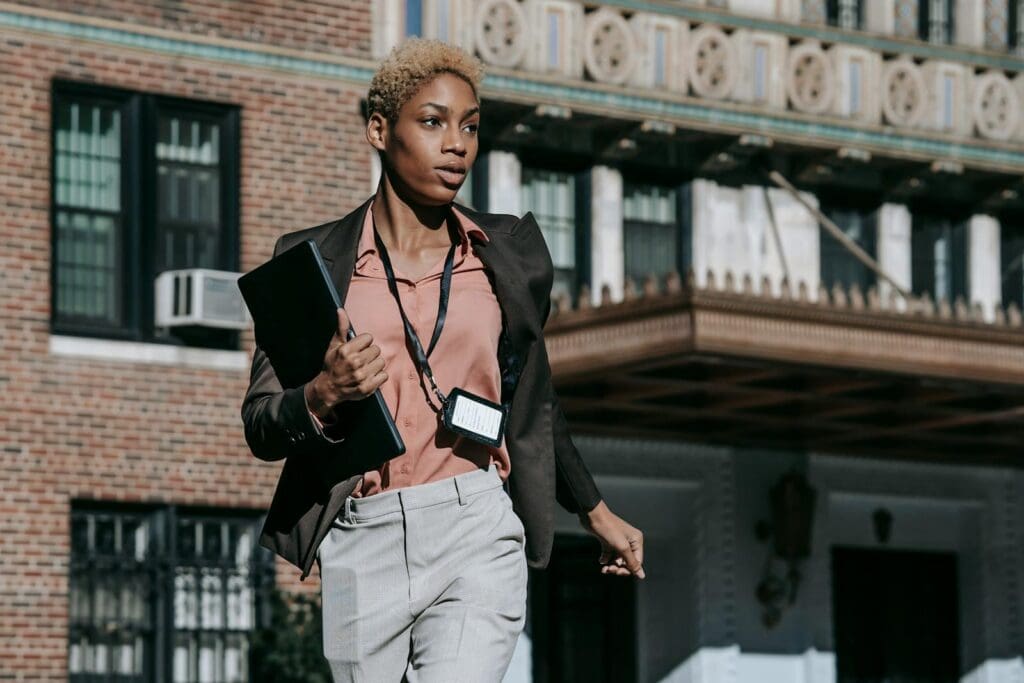 Confident African American female entrepreneur in classy outfit with badge carrying laptop while running fast on street