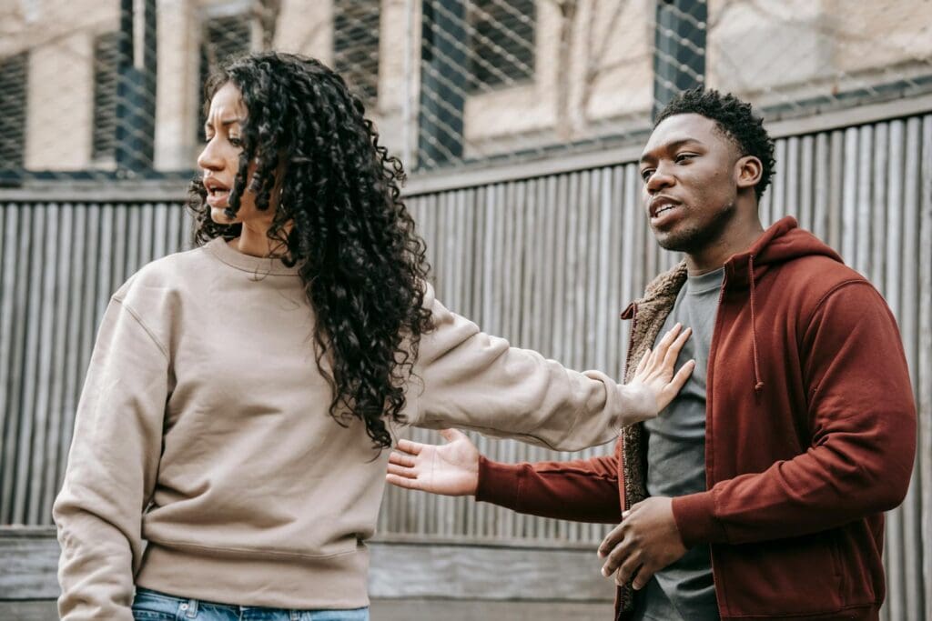 A couple in casual clothing appears to be in a disagreement outdoors with grey fencing background.