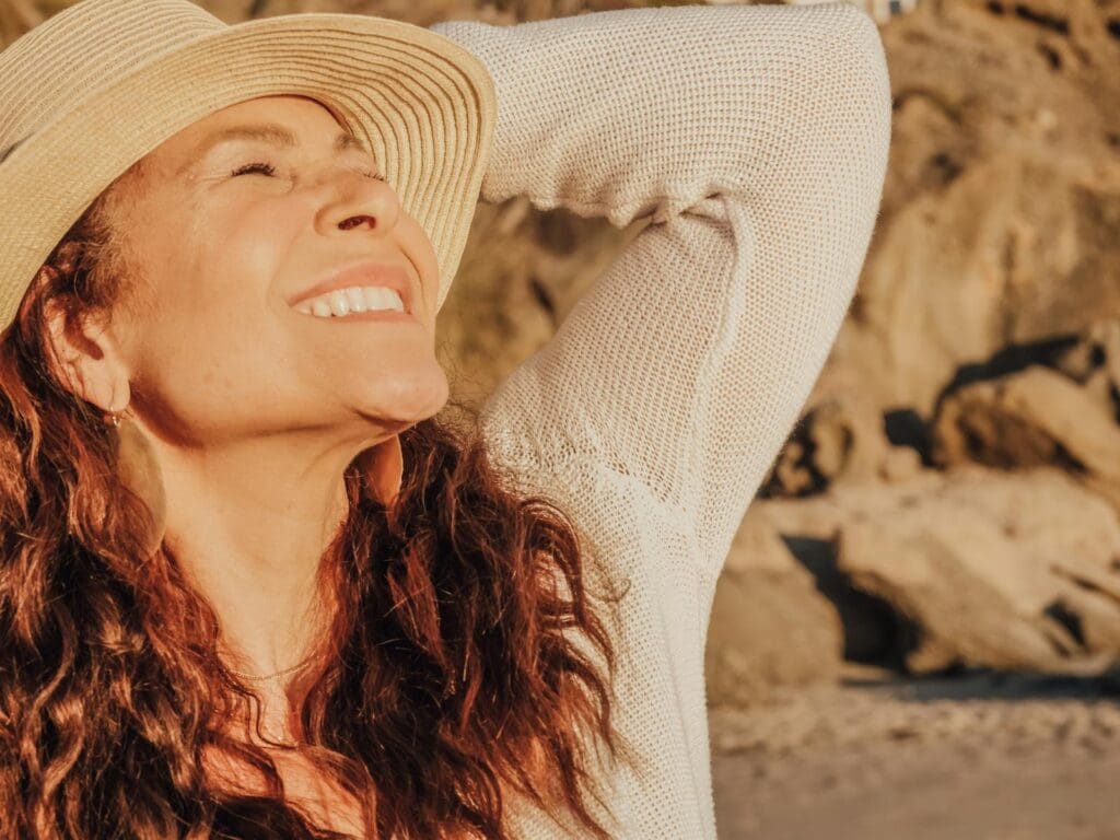 A joyful woman with a hat basks in the sunlight by the seaside, symbolizing relaxation and happiness.