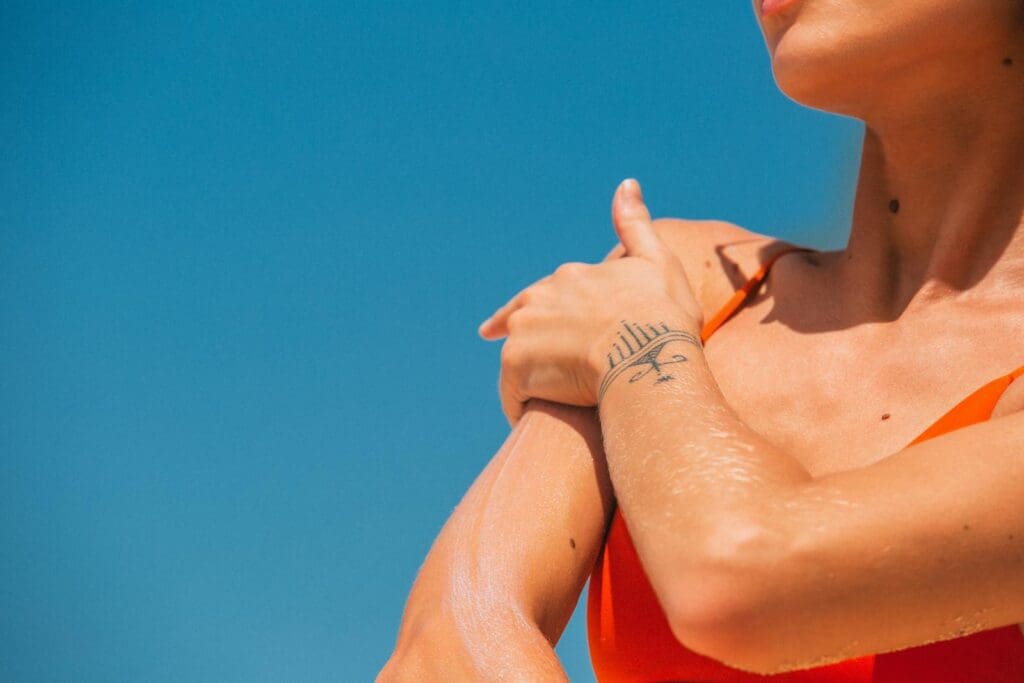 Close-up of a woman applying sunscreen to her shoulder with a tattoo visible in a bright, sunny setting.