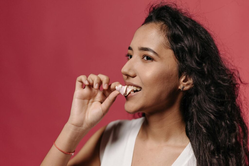 Portrait of a young woman with curly hair biting a candy against a pink background.