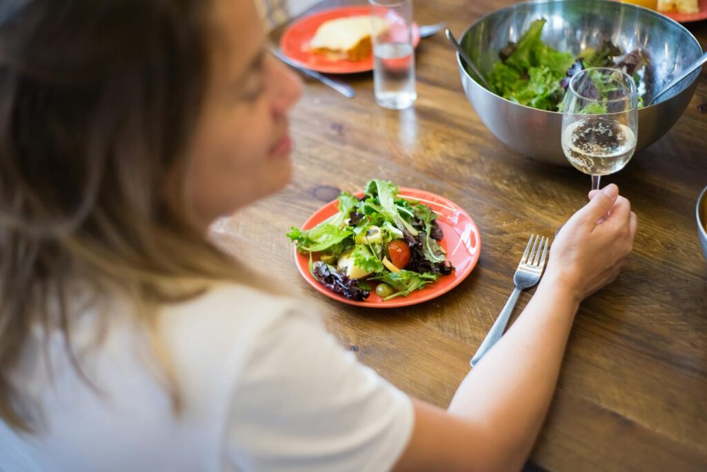 A woman sitting at a wooden table, savoring a fresh vegetable salad with a glass of white wine indoors.