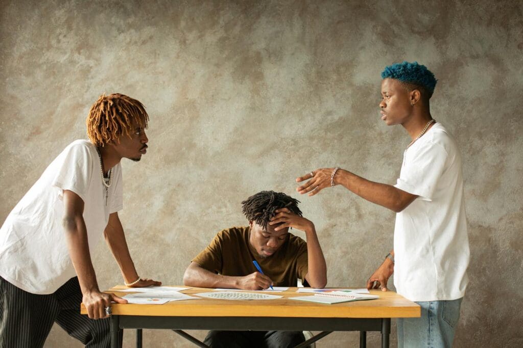 Three men in heated discussion around a table with documents, expressing emotions and ideas.