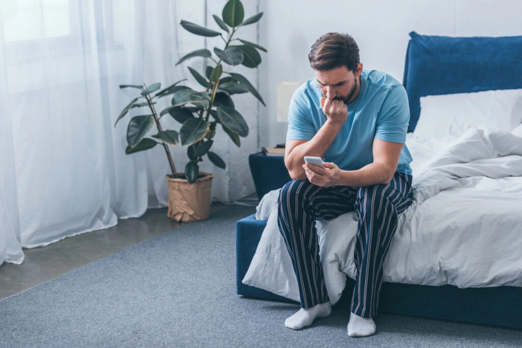 grieving man sitting on bed, covering mouth with hand and using smartphone at home