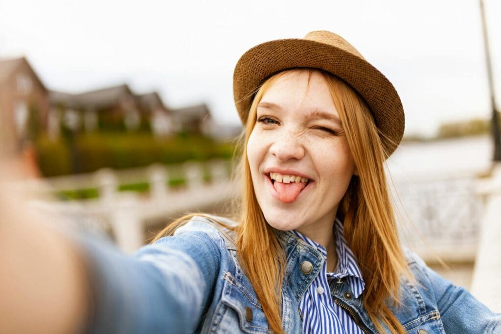 Cheerful woman in denim and fedora takes a playful selfie on a sunny day.