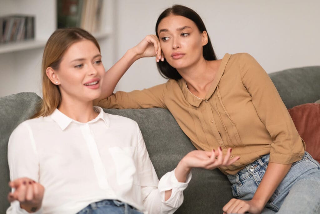 Jealous Woman Listening With Envy To Her Friend Bragging About Great Life Sitting On Sofa In Modern Living Room At Home. Envious Lousy Friends, Fake Friendship Concept. Selective Focus