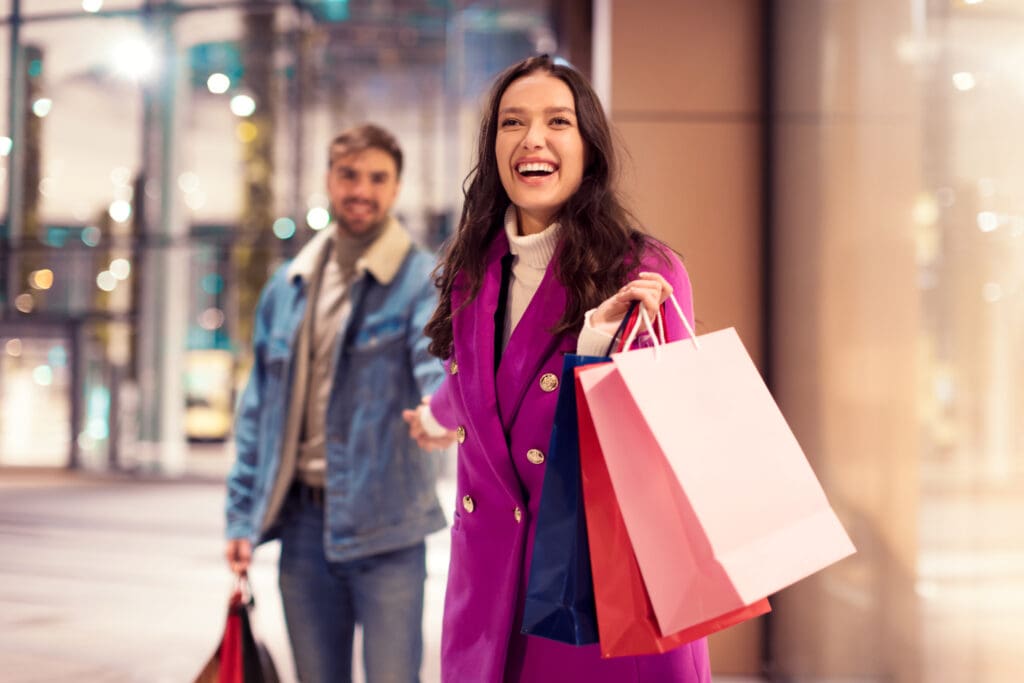 Joyful spouses walking near modern mall outdoor, holding hands and laughing with shopping bags, enjoying winter evening of holiday shopping, epitome of New Year festive consumerism in the city