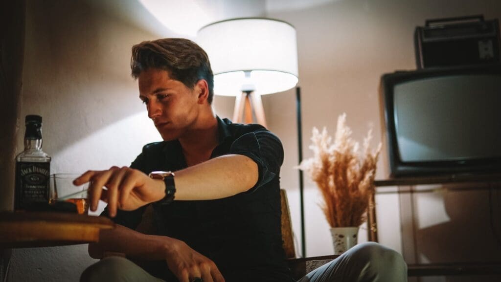 A man in deep thought sits alone with a glass of whiskey in a cozy vintage room.