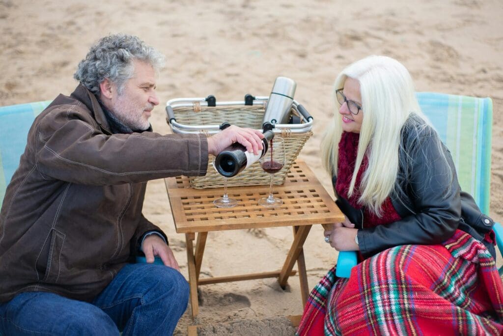 A senior couple enjoys a cozy beach picnic with wine on a wooden table.