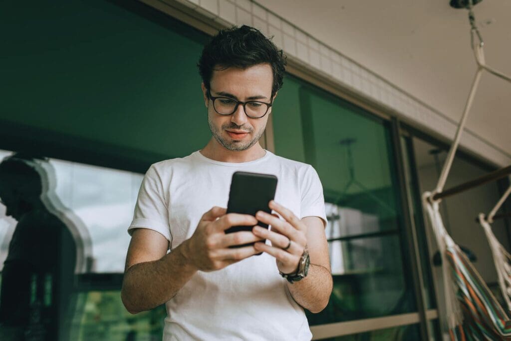 Young man standing on a balcony, using a smartphone, reflecting in the window.