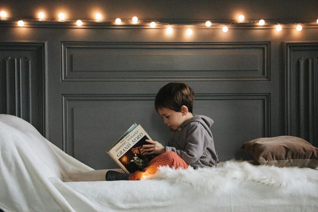 A young boy reads a book in a cozy bedroom setting with warm string lights and soft textures.