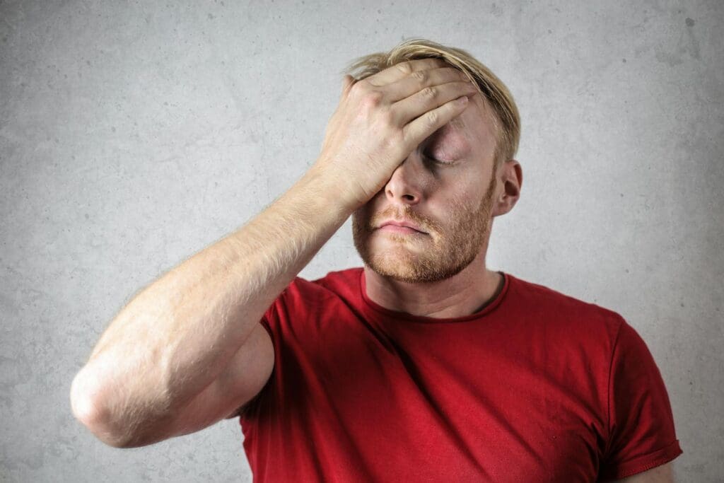 A frustrated man in a red shirt holds his head in stress against a neutral background.