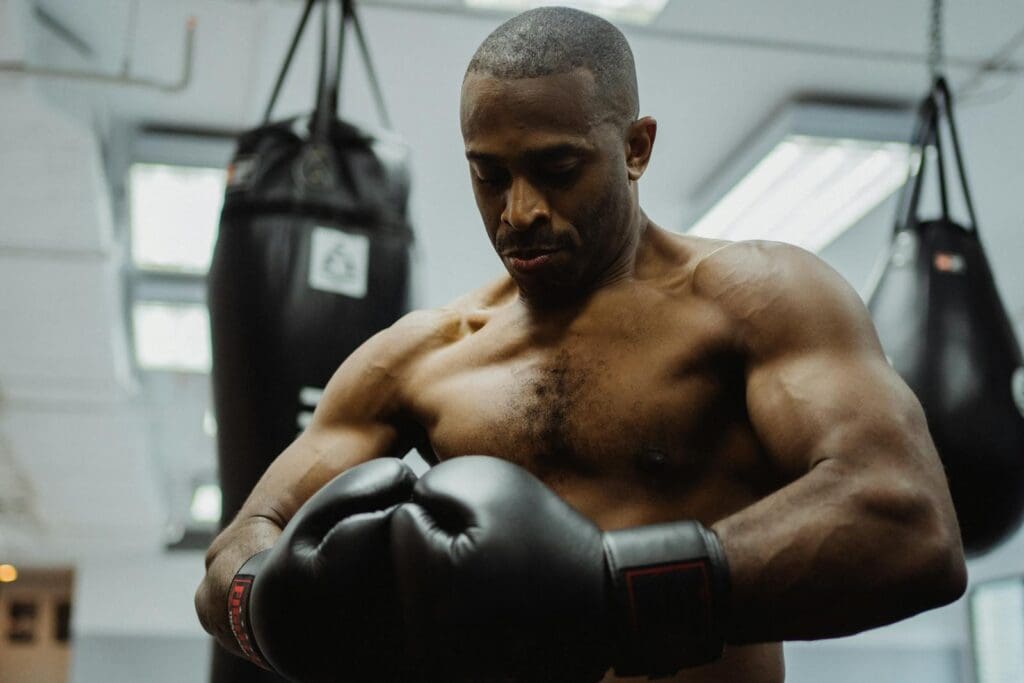 Shirtless black man tightening boxing gloves in gym with punching bags.