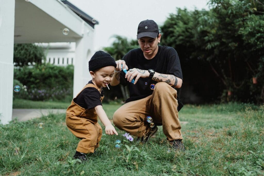 A playful moment between a father and son blowing bubbles outdoors on a sunny day.