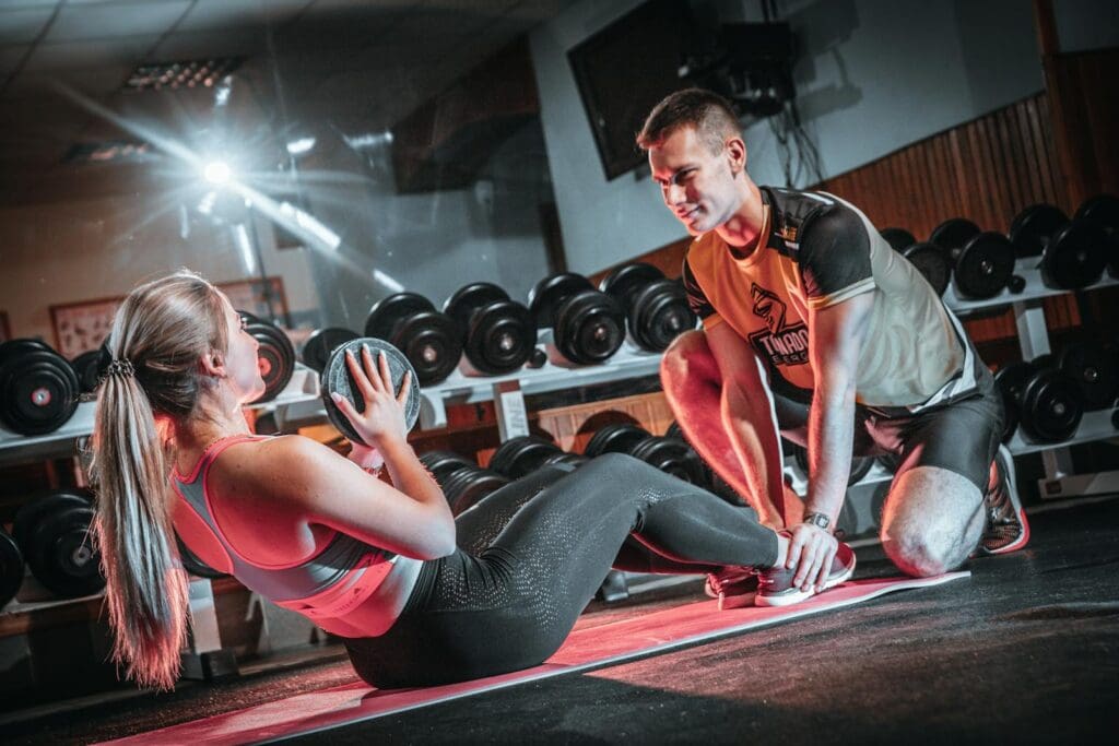A personal trainer assists a woman doing sit-ups with a medicine ball in a gym setting.