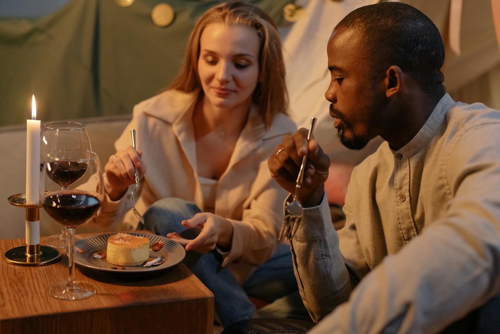 A couple enjoys a romantic candlelit dinner indoors, sharing wine and dessert.