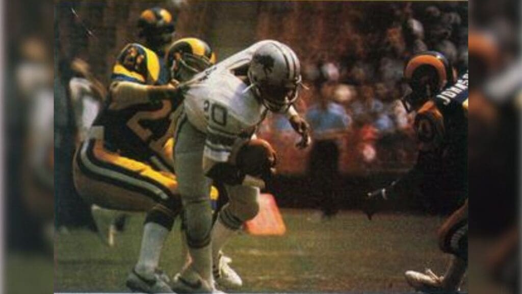 football card from the 1986 Jeno's Pizza NFL football card stickers set of Detroit Lions running back Billy Sims rushing the ball against the Los Angeles Rams during an away game on September 7, 1980 at Anaheim Stadium.