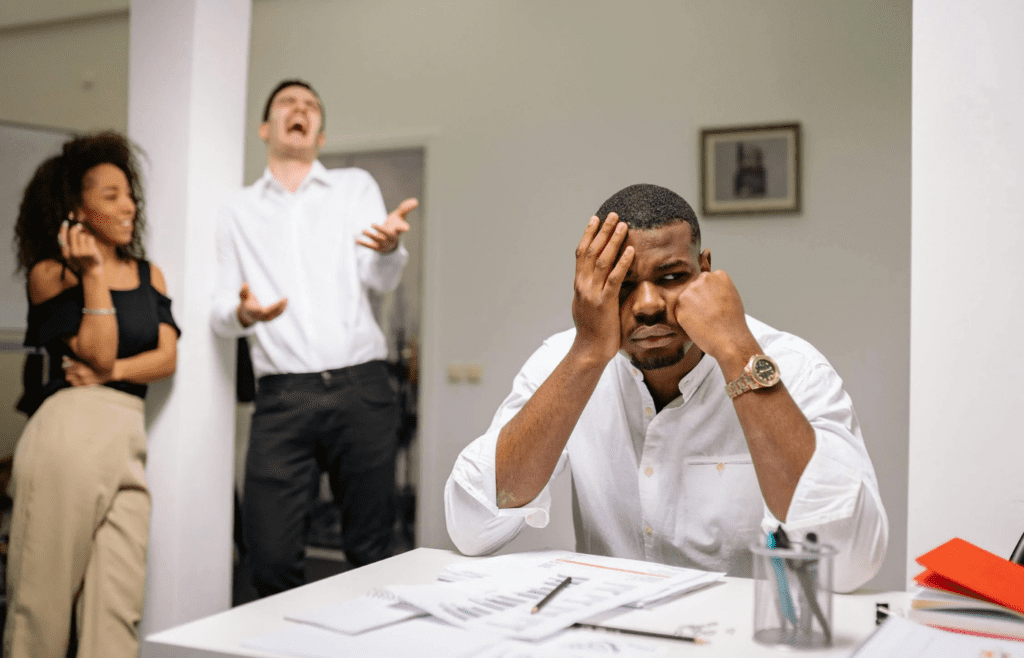Frustrated man at desk while coworkers laugh