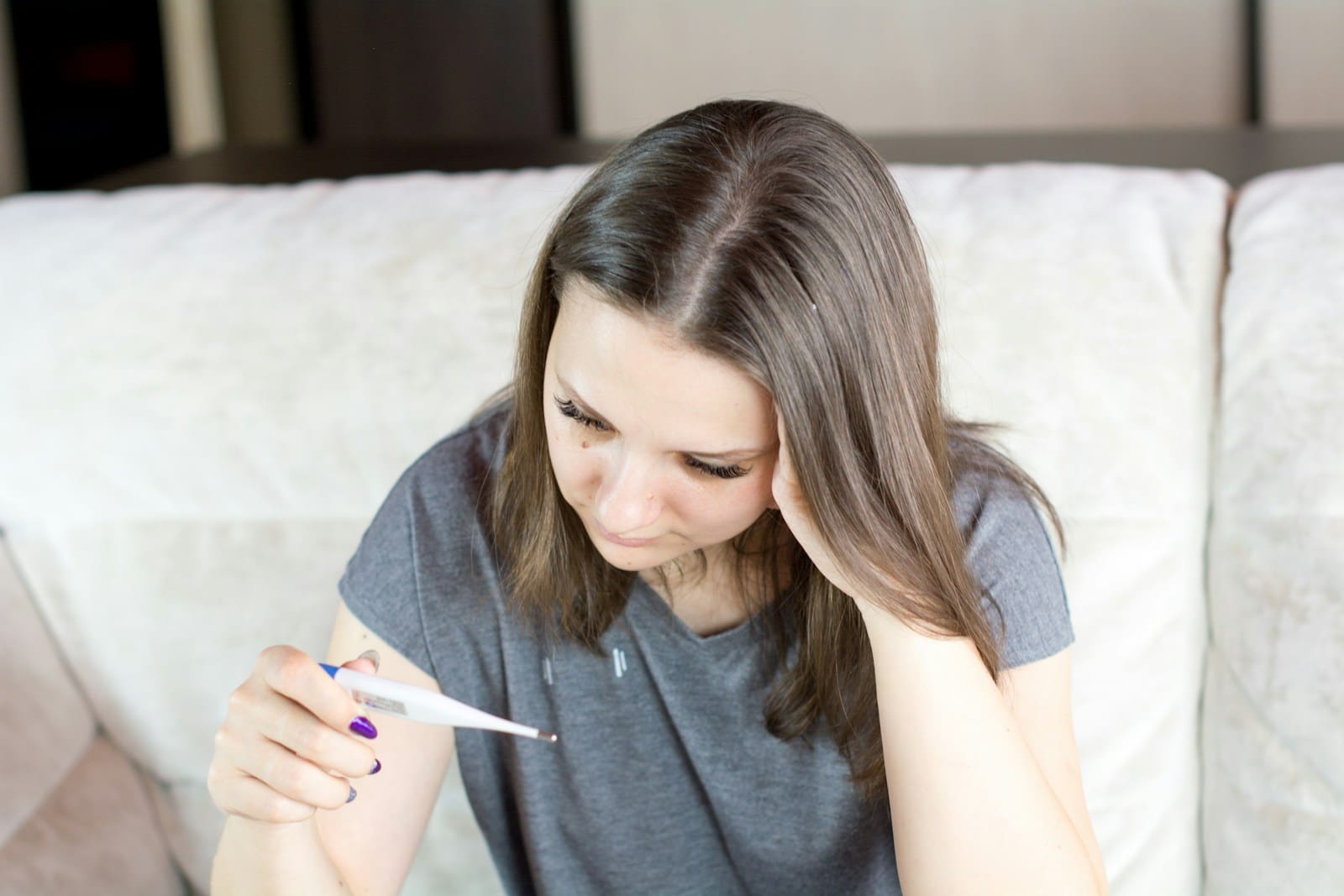 a sick woman sitting on a couch holding a thermometer
