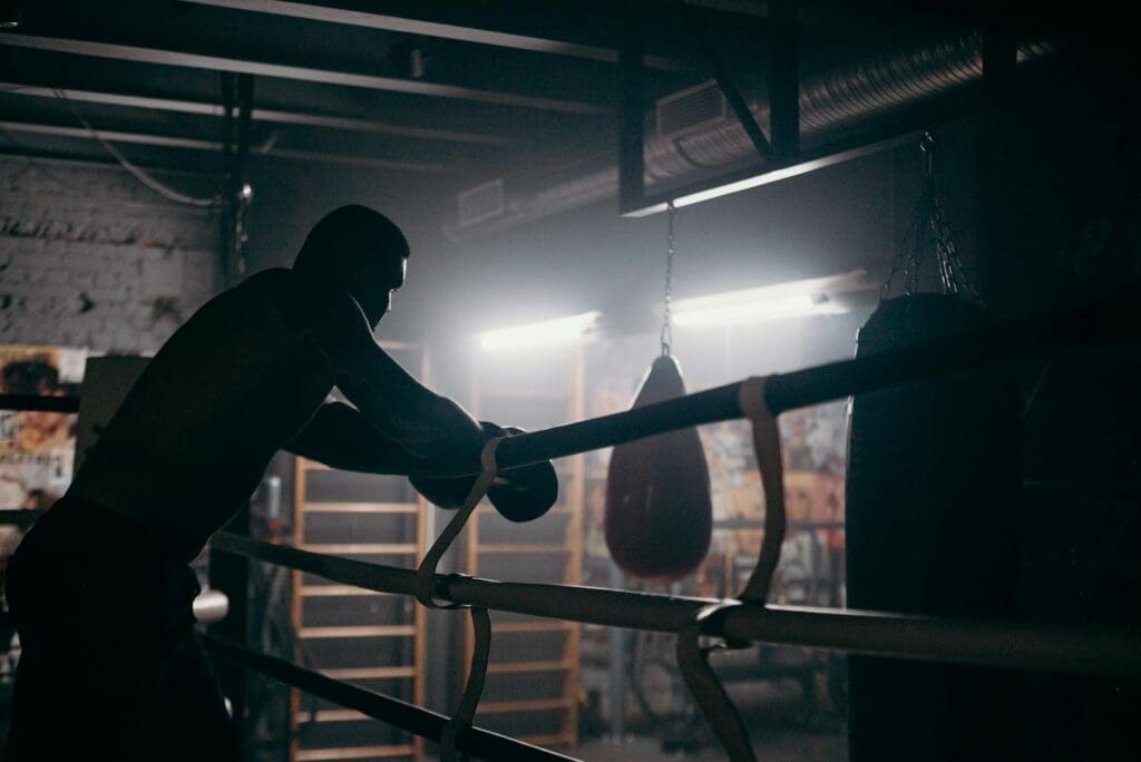 Moody silhouette of a boxer leaning in a dimly lit gym with punching bag and boxing ring.